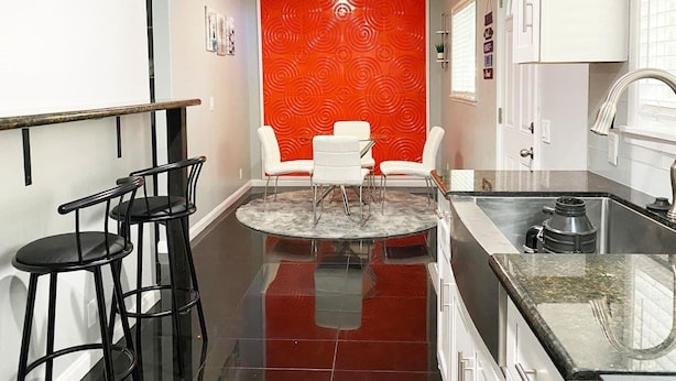 Una cocina moderna con encimera negra, armarios blancos y una pared de acento roja. Hay dos sillas blancas y una mesa redonda en el centro.