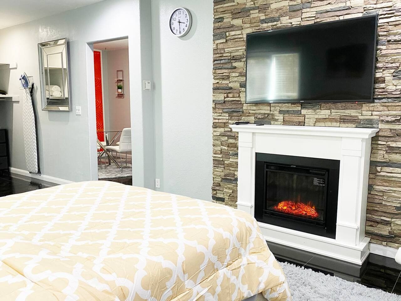 A bedroom with a fireplace, a bed with a patterned bedspread, a clock on the wall, and a television mounted above the fireplace.