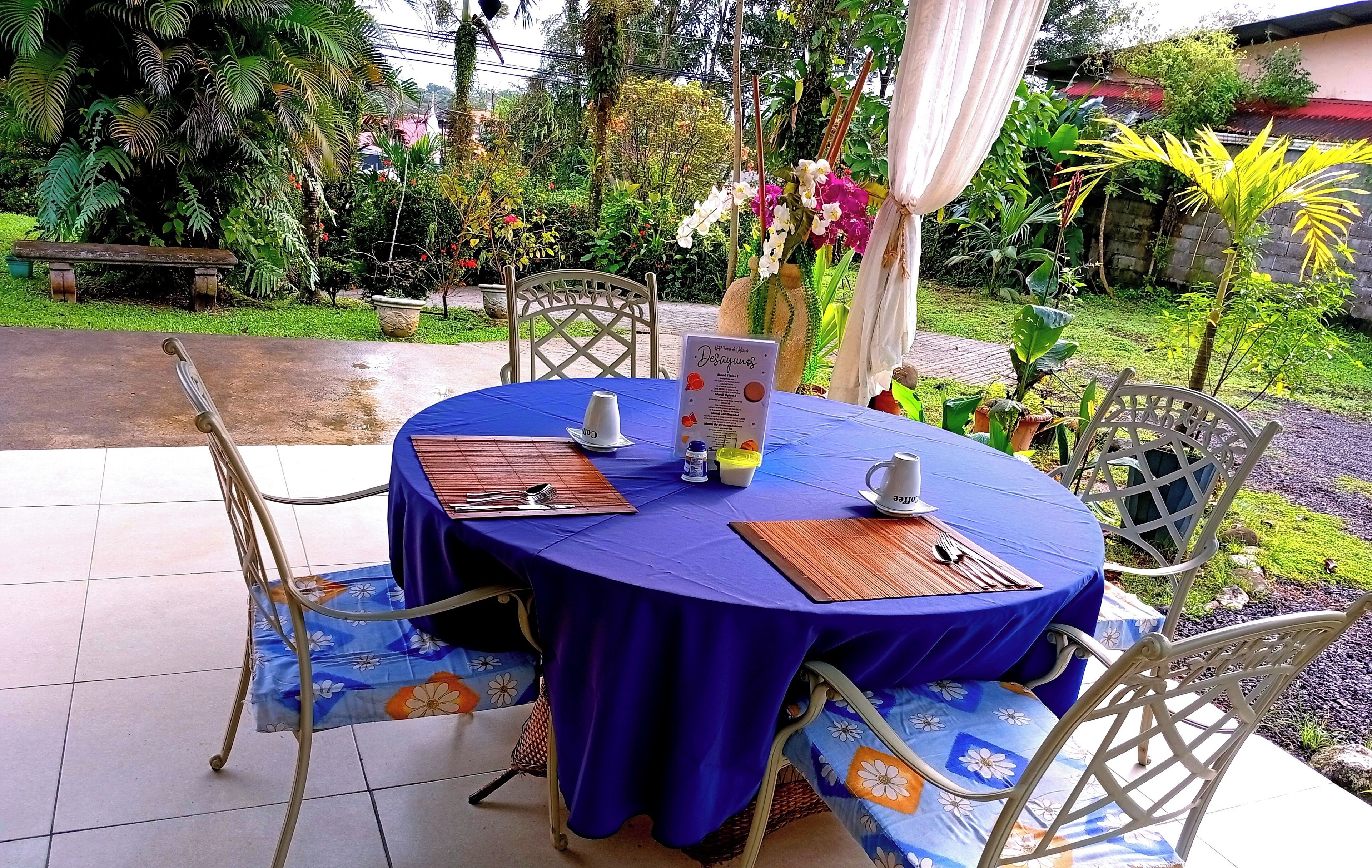 An outdoor dining area with round tables covered in blue tablecloths, metal chairs with floral cushions, and a menu stand.