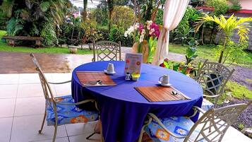 An outdoor dining area with round tables covered in blue tablecloths, metal chairs with floral cushions, and a menu stand.