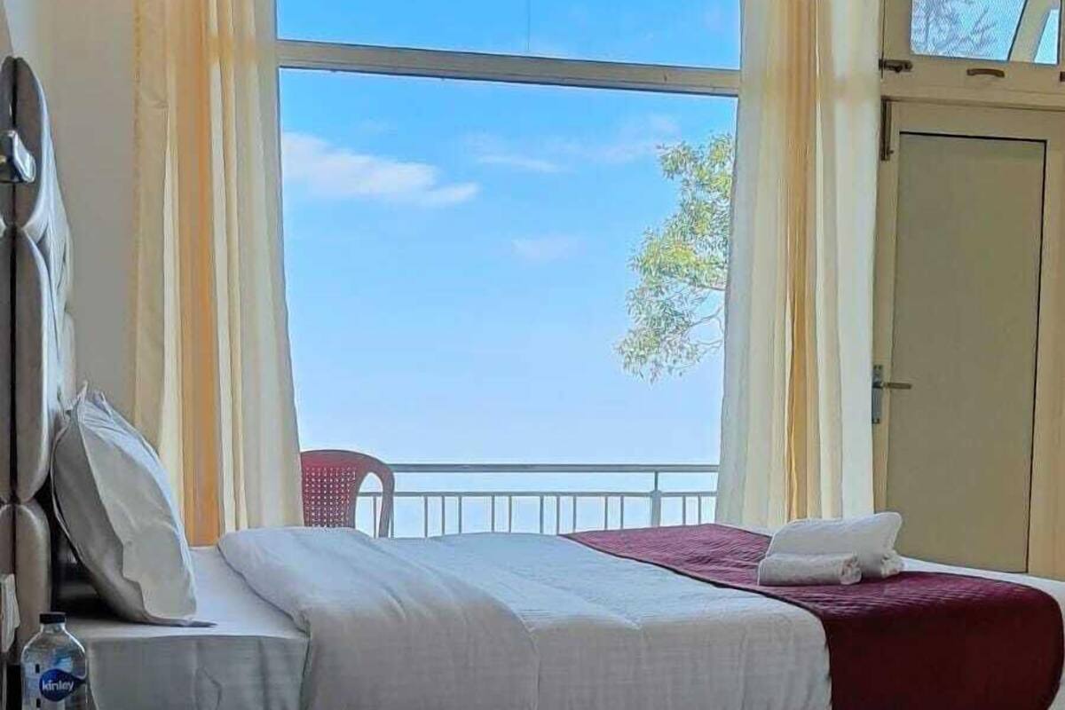 A hotel room with a bed, a nightstand, a water bottle, and a view of the sky through large windows.