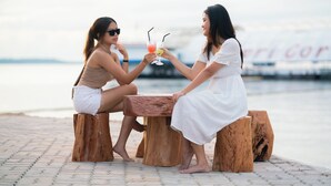 Two women sitting at a wooden table outdoors with drinks, near a body of water and a sign in the background. - Kepri Coral Resort (Galang)