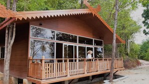 A wooden cabin with a large window, a balcony, and a sandy ground. - Kepri Coral Resort (Galang)