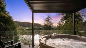 A hot tub on a wooden deck with a glass railing, overlooking a lake and forested hills.