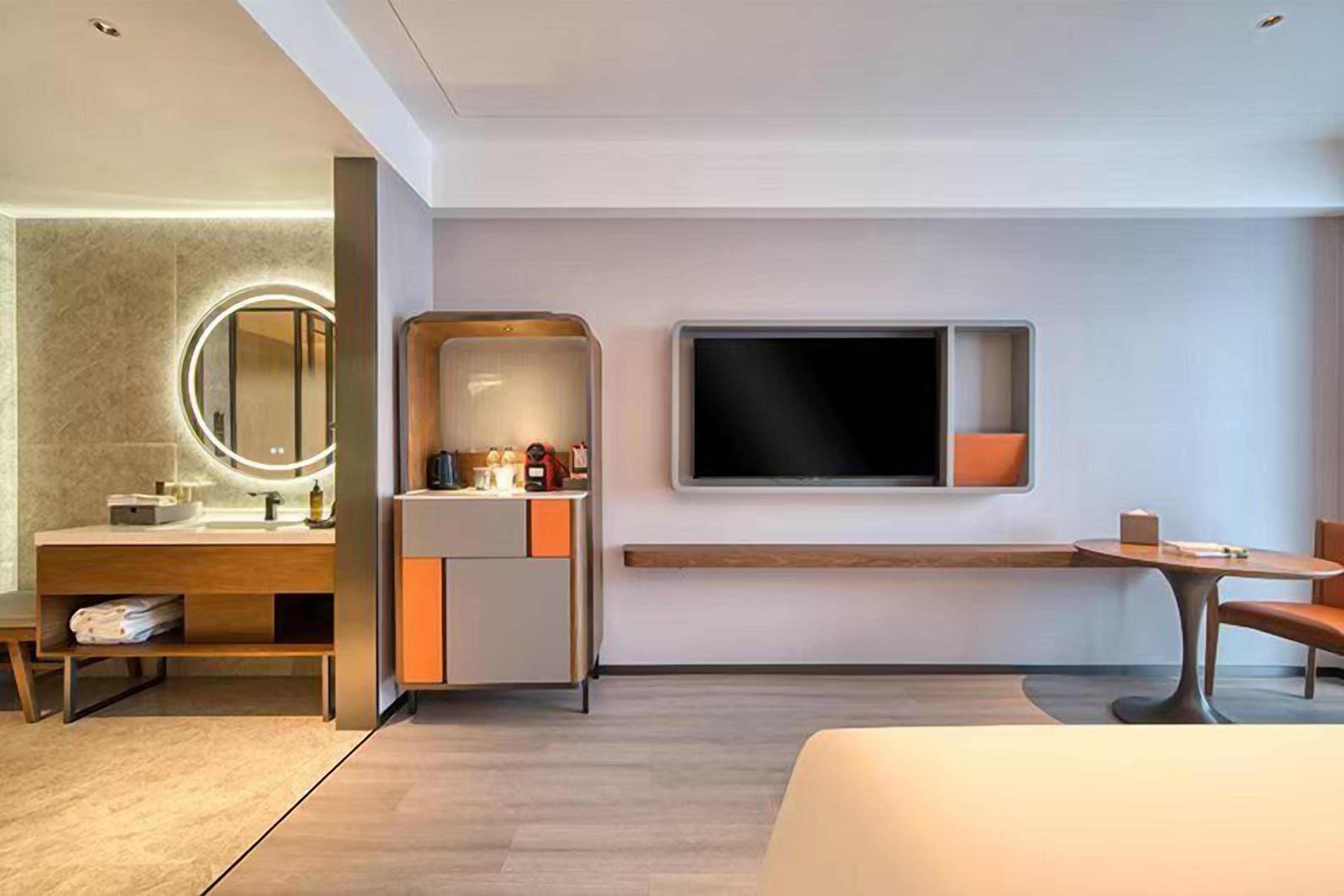 A modern hotel room with a wooden vanity, a large mirror, a flat-screen TV, a small table, and a comfortable bed.