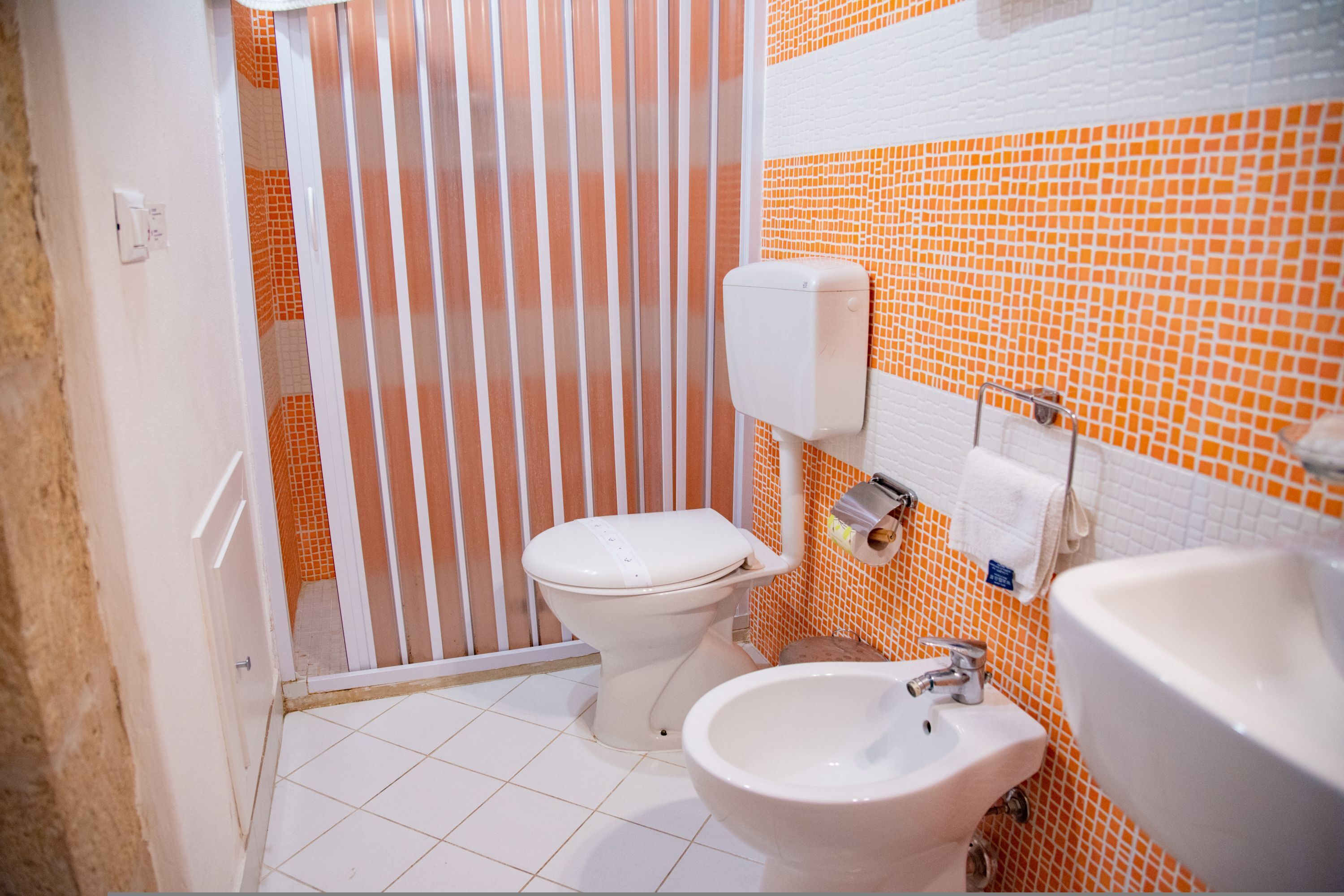 A bathroom with a toilet, bidet, and sink, featuring orange and white tiled walls and a white floor.
