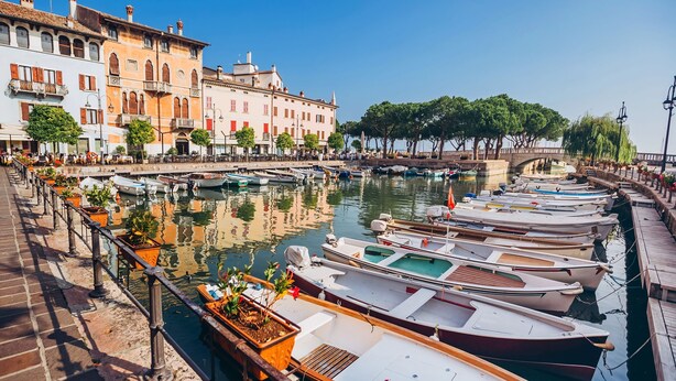 Un port de plaisance où les bateaux sont amarrés le long d’une promenade en bord de mer, avec des bâtiments historiques et des arbres.