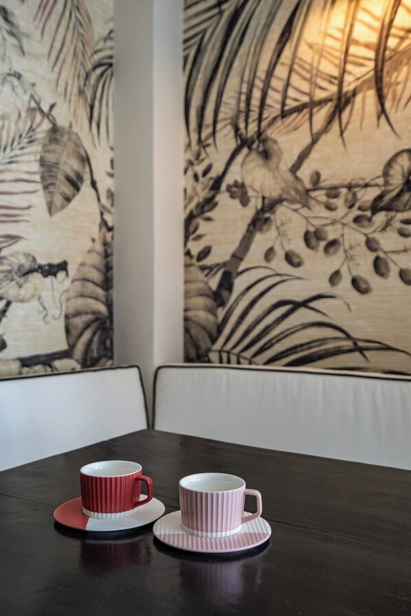 A dining area with two red and white cups on a dark wooden table, white cushioned seating, and a wall with a bird and plant mural.