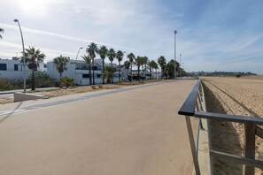 A coastal walkway with a sandy beach, palm trees, and modern buildings in the background.