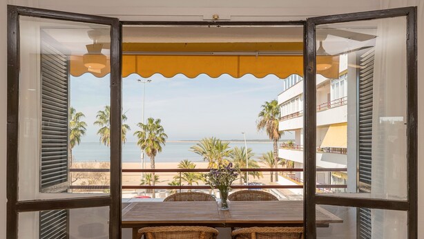 A balcony with wicker chairs, a table with a vase of flowers, and a view of the beach and palm trees. - Ventana A Do ana in Sanlucar de Barrameda (Sanlucar de Barrameda)