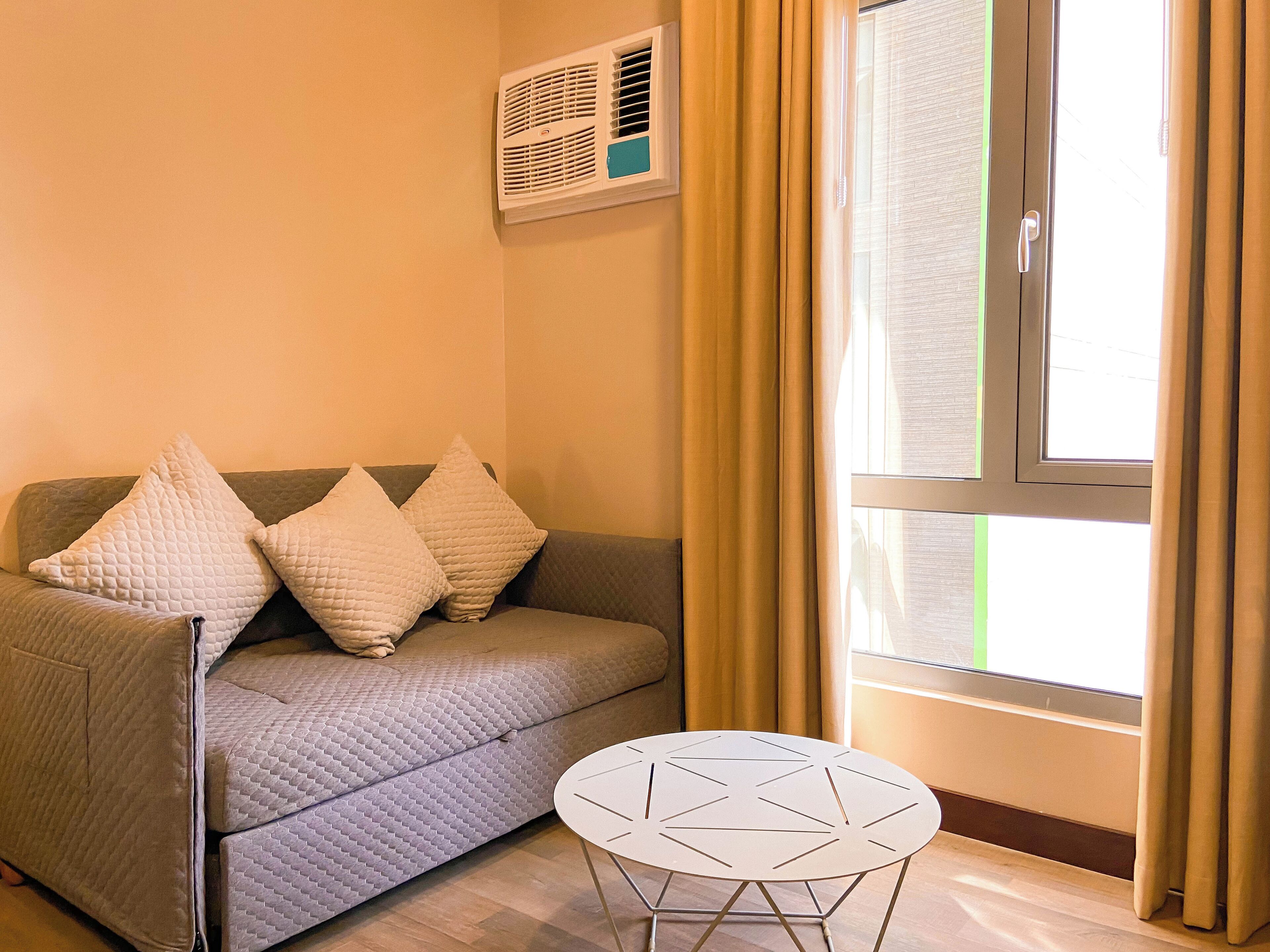 Una sala de estar con un sofá, tres almohadas, una mesita redonda y una ventana con cortinas.