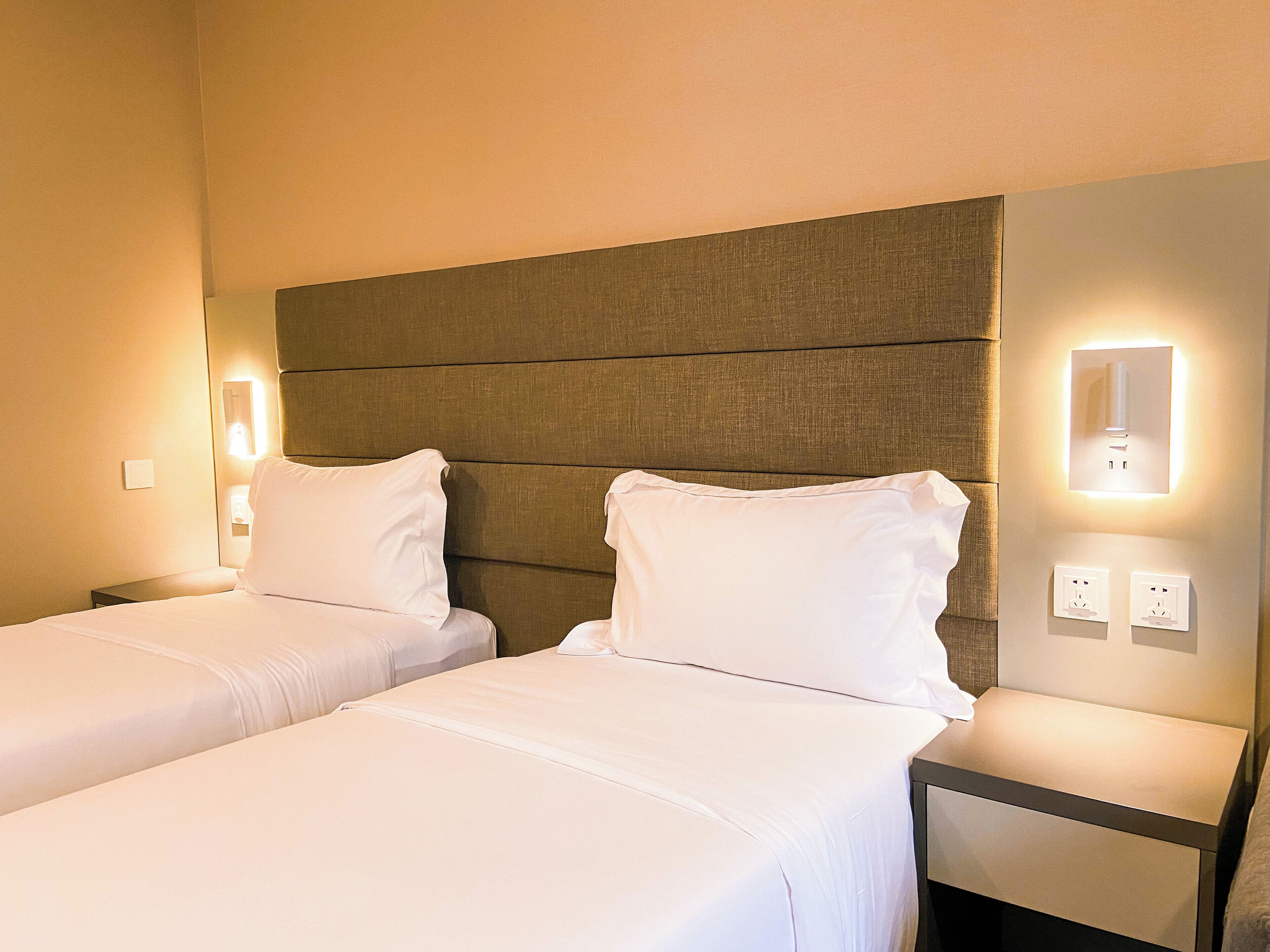 A hotel room with two single beds, a headboard, bedside lamps, and a nightstand with a lamp.