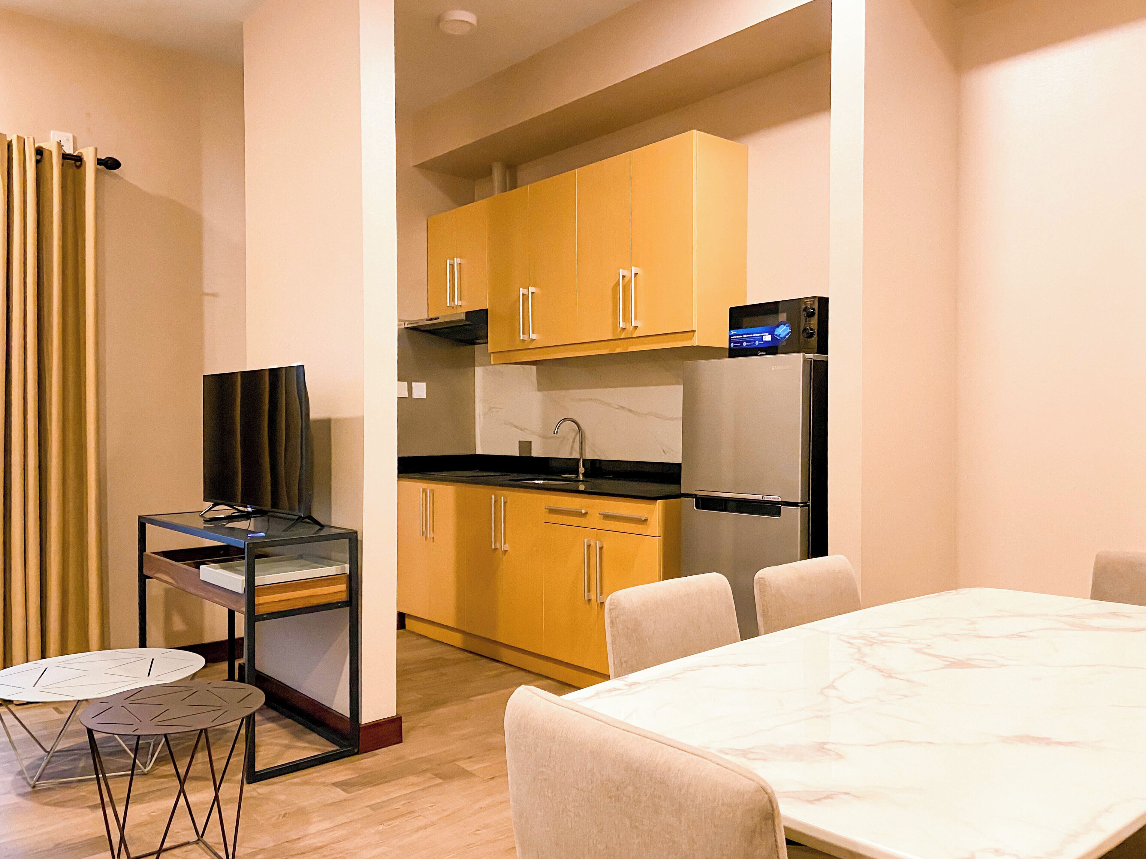 A modern kitchen with wooden cabinets, a stainless steel refrigerator, and a built-in oven. There is a dining area with a marble table and chairs.
