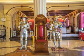 Deux statues représentant des personnages en armure se trouvent dans un hall d’entrée orné d’un grand lustre et doté d’un siège rembourré de couleur rouge.