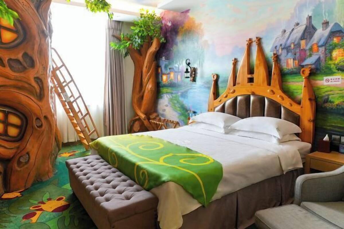 Une chambre à coucher avec un grand lit, une tête de lit en bois, une couvre-lit verte, un banc et une fresque représentant un village sur le mur.