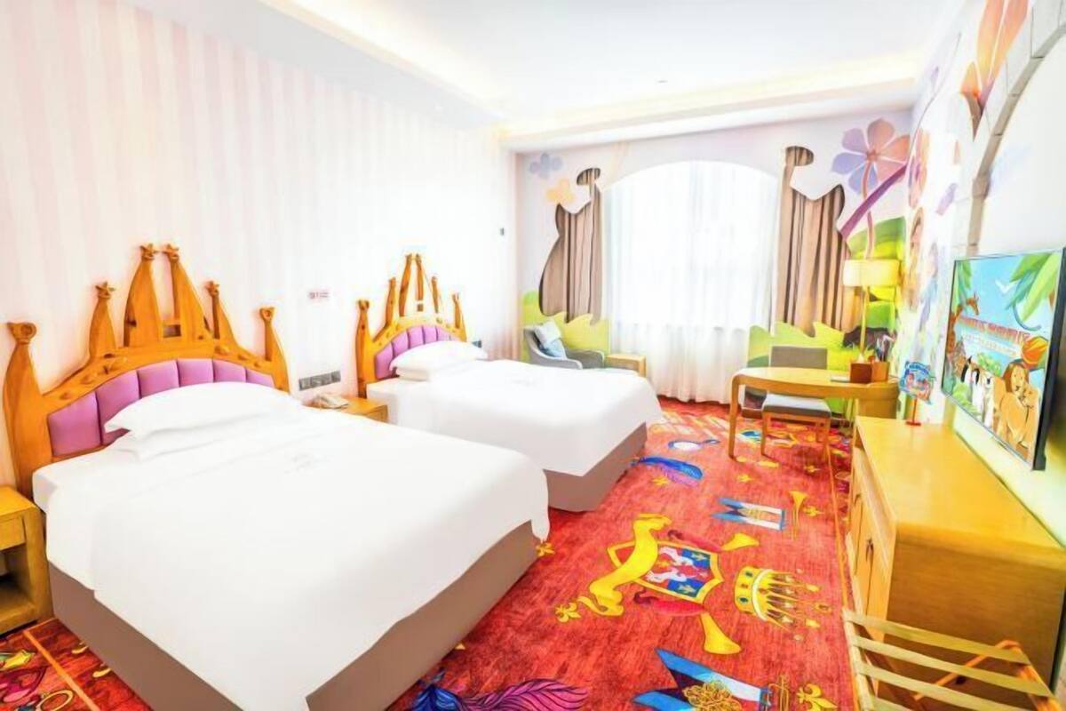 Une chambre d’hôtel avec deux lits, une grande fenêtre avec des rideaux, une moquette colorée et un bureau avec une lampe.