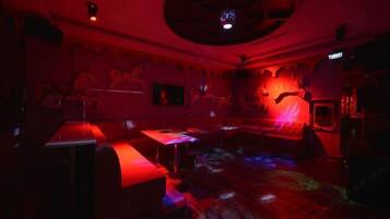 Une pièce faiblement éclairée, baignée d’une lumière rouge, avec un ventilateur de plafond et un coin bar équipé de tabourets.