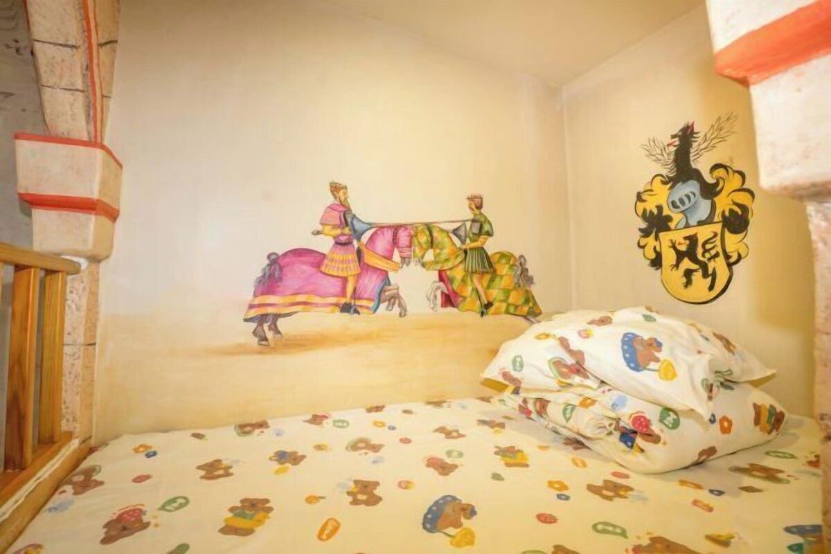 Ein Kinderzimmer mit Wandgemälde von zwei Frauen auf einem Pferd, einem Bett mit Teddybär-Muster und einem Wappen an der Wand.