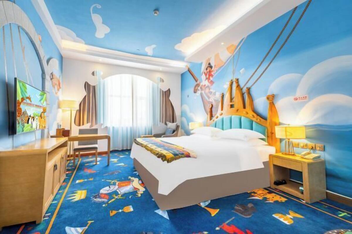 Une chambre d’hôtel avec un grand lit, un bureau et une chaise. Il y a une fenêtre avec des rideaux, une fresque murale et un tapis orné de personnages de dessins animés.