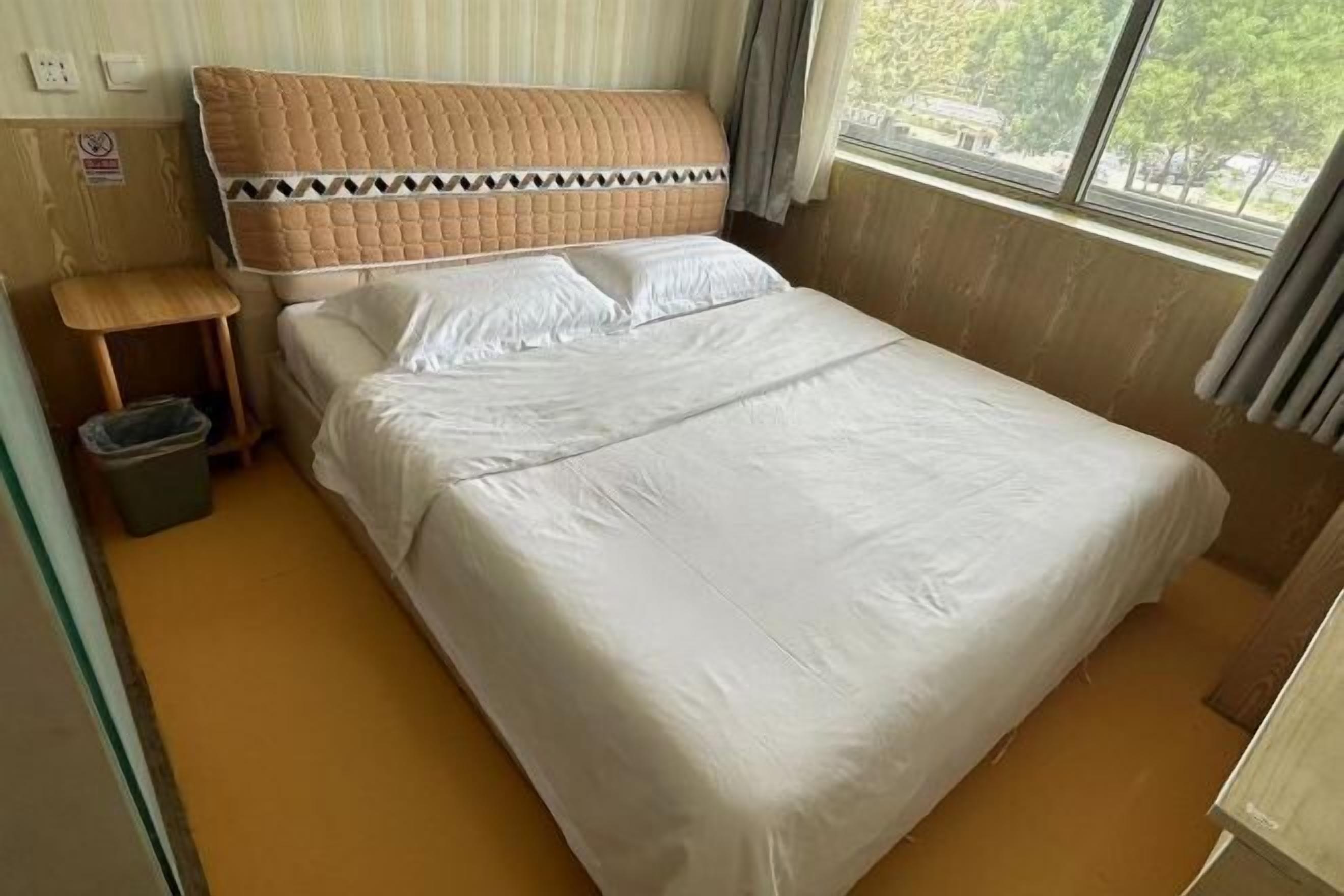 Una cama bien hecha con cabecera, una mesita de noche pequeña y un cubo de basura.