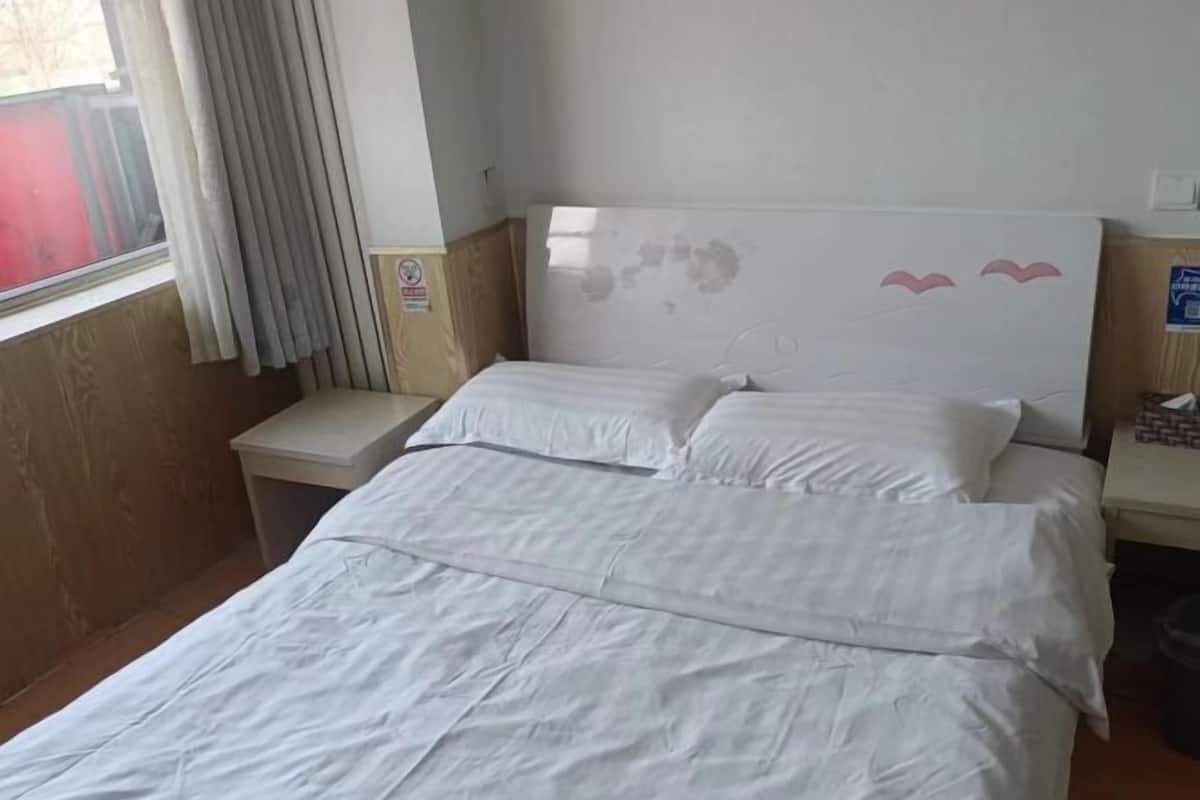 A hotel room with a bed, a nightstand, a window with curtains, and a wall-mounted sign.