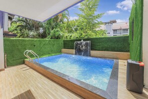 A swimming pool with a waterfall feature, surrounded by green hedges and a wooden deck. - 3BHK w/Pool - Dream Habitat by Universe Villas (Mawal)