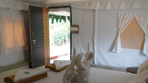 Un lit simple avec un matelas blanc, une serviette pliée et une serviette enroulée posées sur le lit. On y trouve une petite table recouverte d’une nappe blanche et sur laquelle se trouve une bouteille. La pièce a un plafond et des murs blancs, ainsi qu’une fenêtre avec des rideaux blancs.