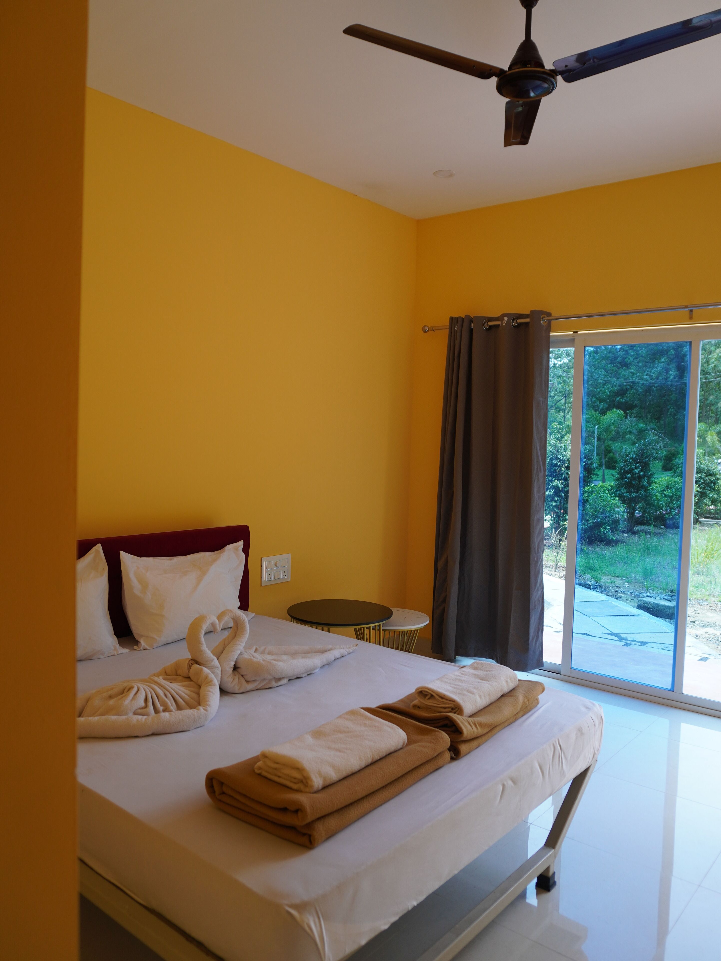 Habitación de hotel con cama, ventilador de techo, ventana con cortinas y vistas a zonas verdes.