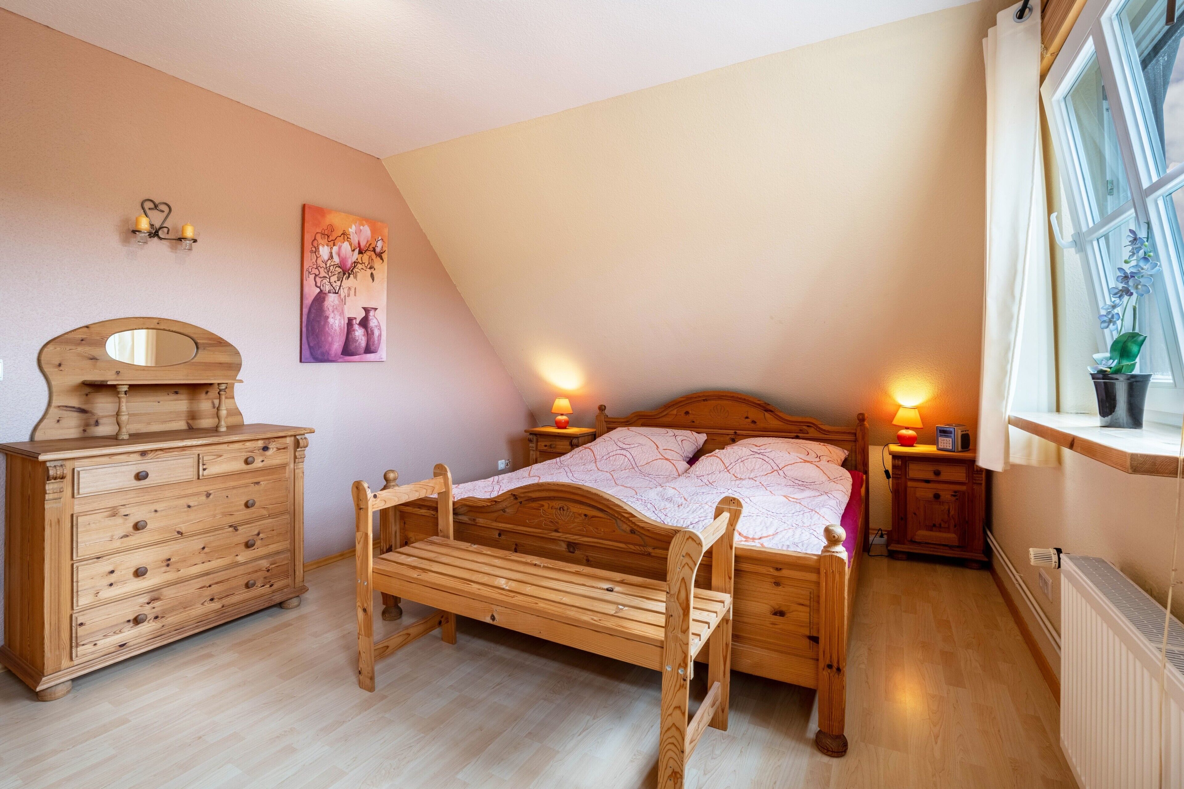 Een slaapkamer met een houten bed, een houten dressoir, een bankje, een nachtkastje met een lamp en een raam met een plant.