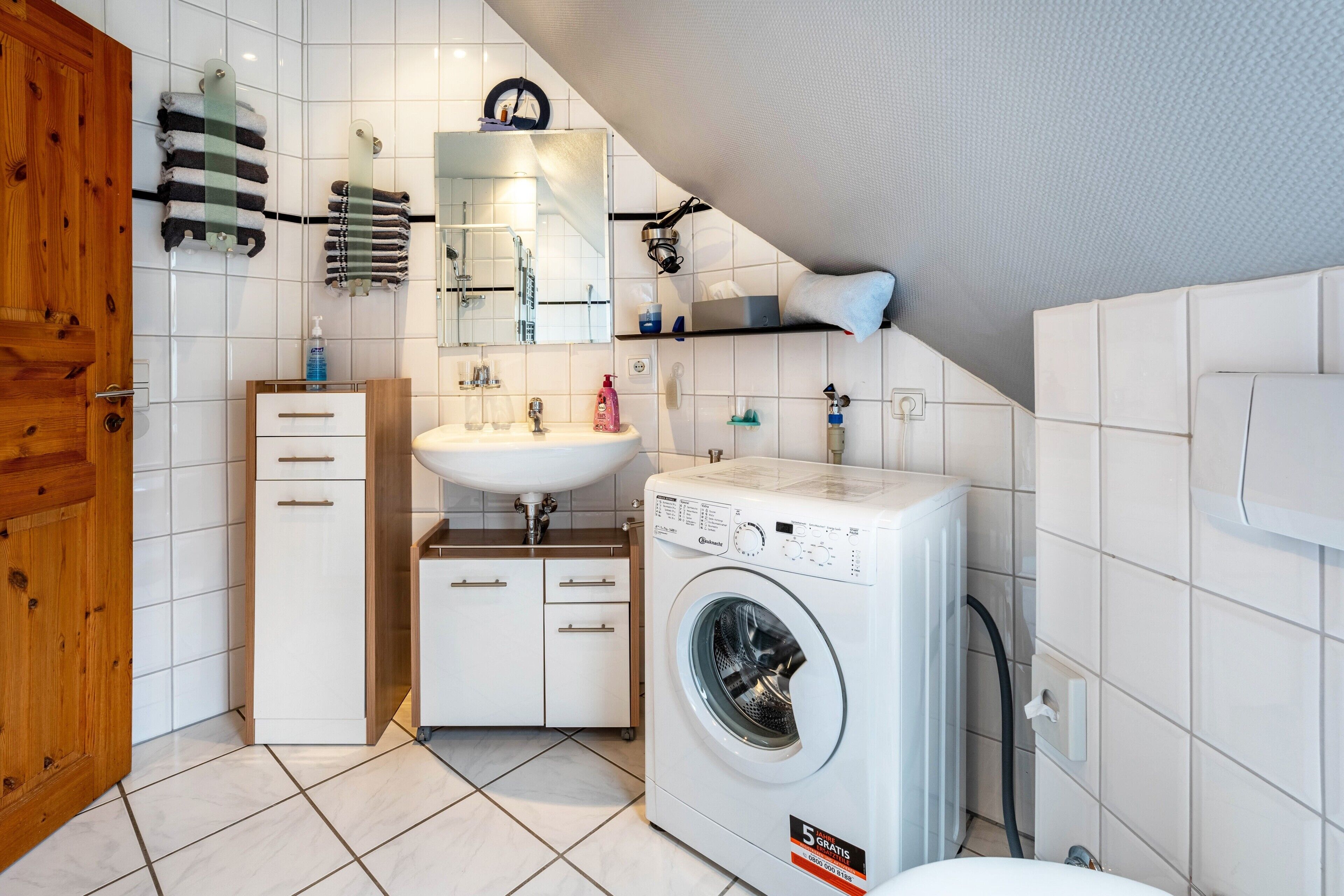A modern bathroom with a sink, mirror, towel rack, wooden door, and a front-loading washing machine.