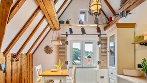 Ein Essbereich mit Holzbalken, einem Tisch mit Stühlen, einem Fenster mit Ausblick und einer Küchenzeile mit Schränken und Heizkörper.