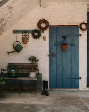 A rustic door with a heart-shaped decoration, a bench, a potted plant, and a watering can against a textured wall.