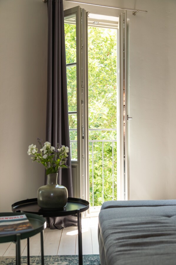 A room with a bed, a small table with a vase, a window with curtains, and a view of greenery outside. - Quiet Boutique Apartment with Courtyard Balcony (Großostheim)