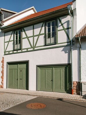 Un bâtiment à deux étages avec des fenêtres et des portes encadrées de vert, un toit recouvert de tuiles et un sol pavé avec un regard d’égout.