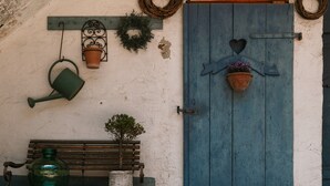 Eine rustikale Tür mit herzförmiger Dekoration, eine Bank, ein Blumentopf und ein Gießkanne vor einer strukturierten Wand.
