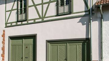 Un bâtiment à deux étages avec des fenêtres et des portes encadrées de vert, un toit recouvert de tuiles et un sol pavé avec un regard d’égout.
