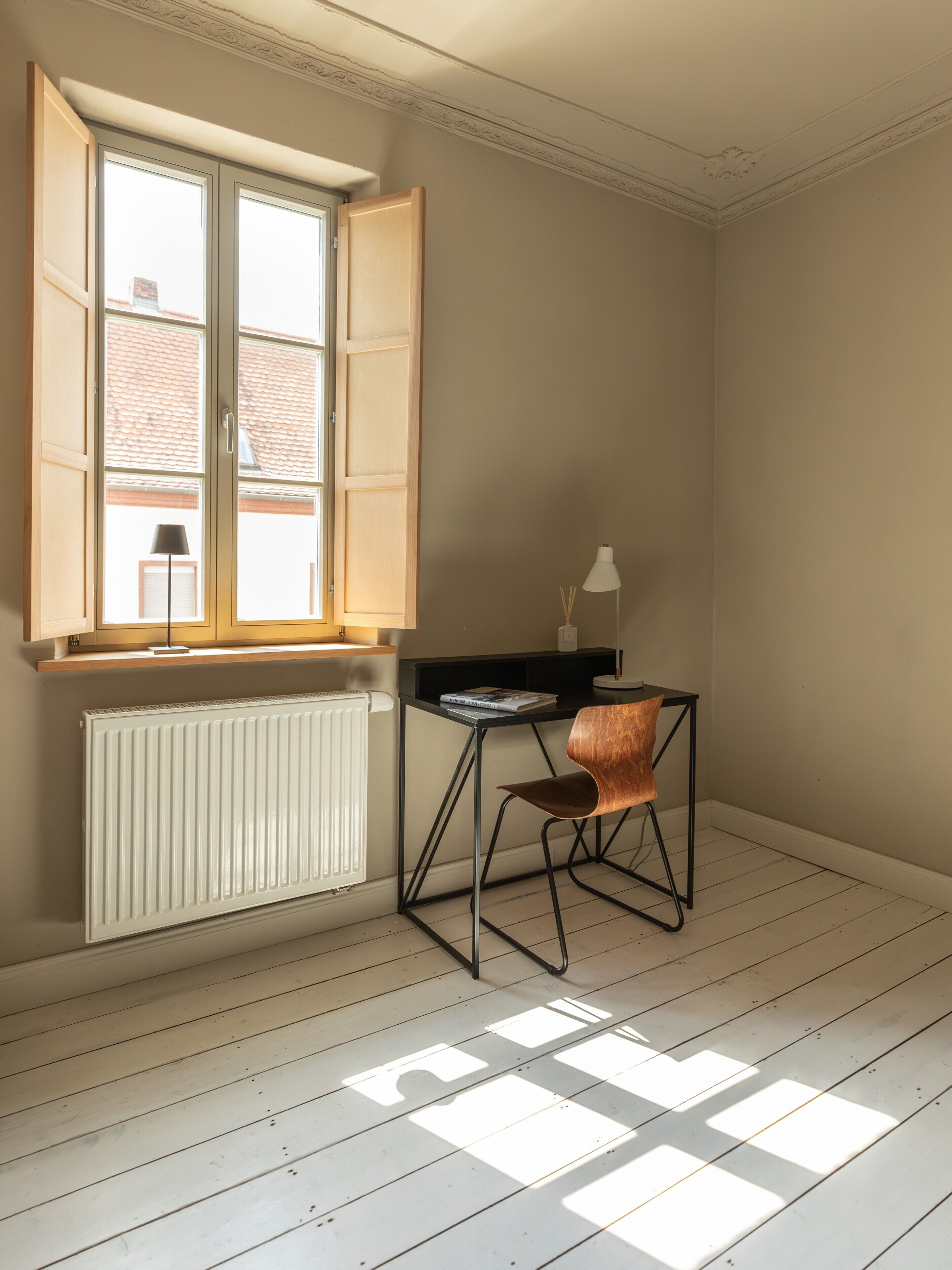 Una habitación con un radiador, un escritorio con silla, una ventana con contraventanas abiertas y una lámpara.