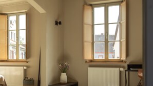 Ein Zimmer mit großem Fenster, Heizkörper, einem gewebten Teppich und einem kleinen Tisch mit einer Vase.