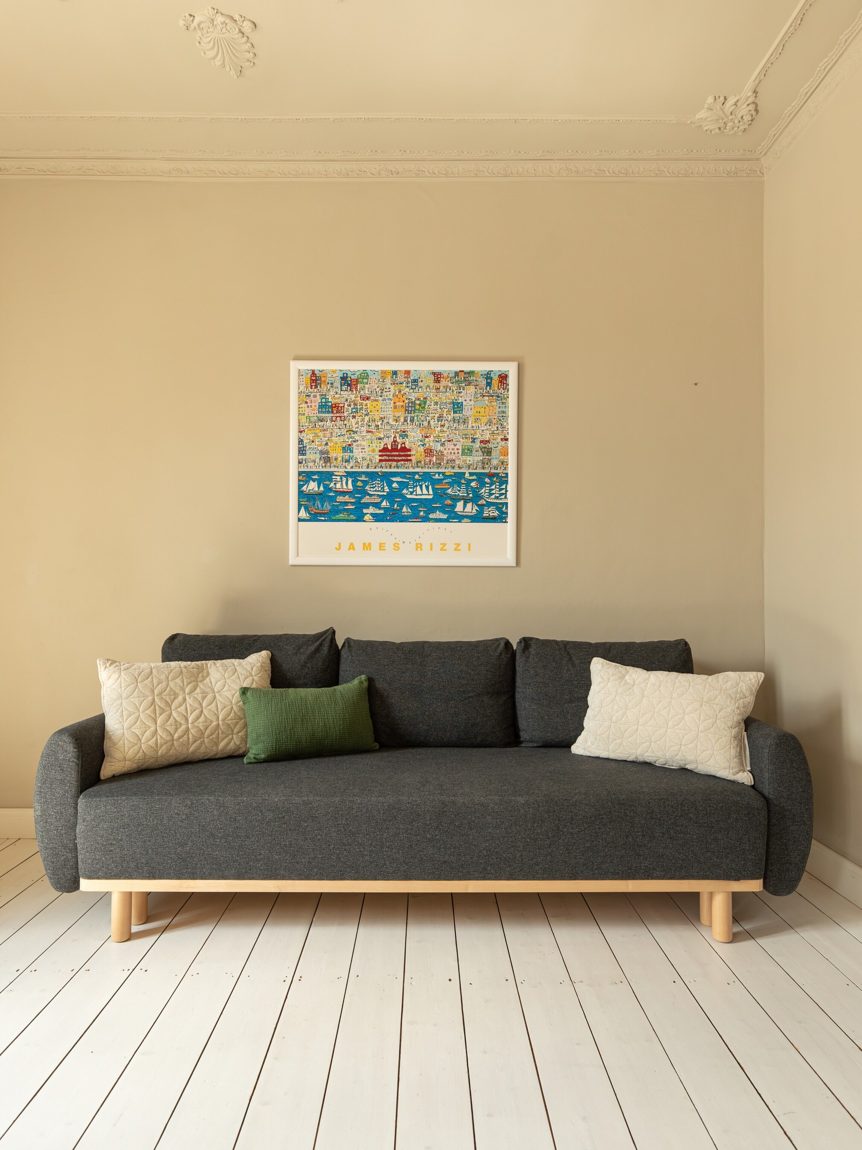 Een grijze bank met houten poten, erboven een ingelijst kunstwerk, en een licht houten vloer.
