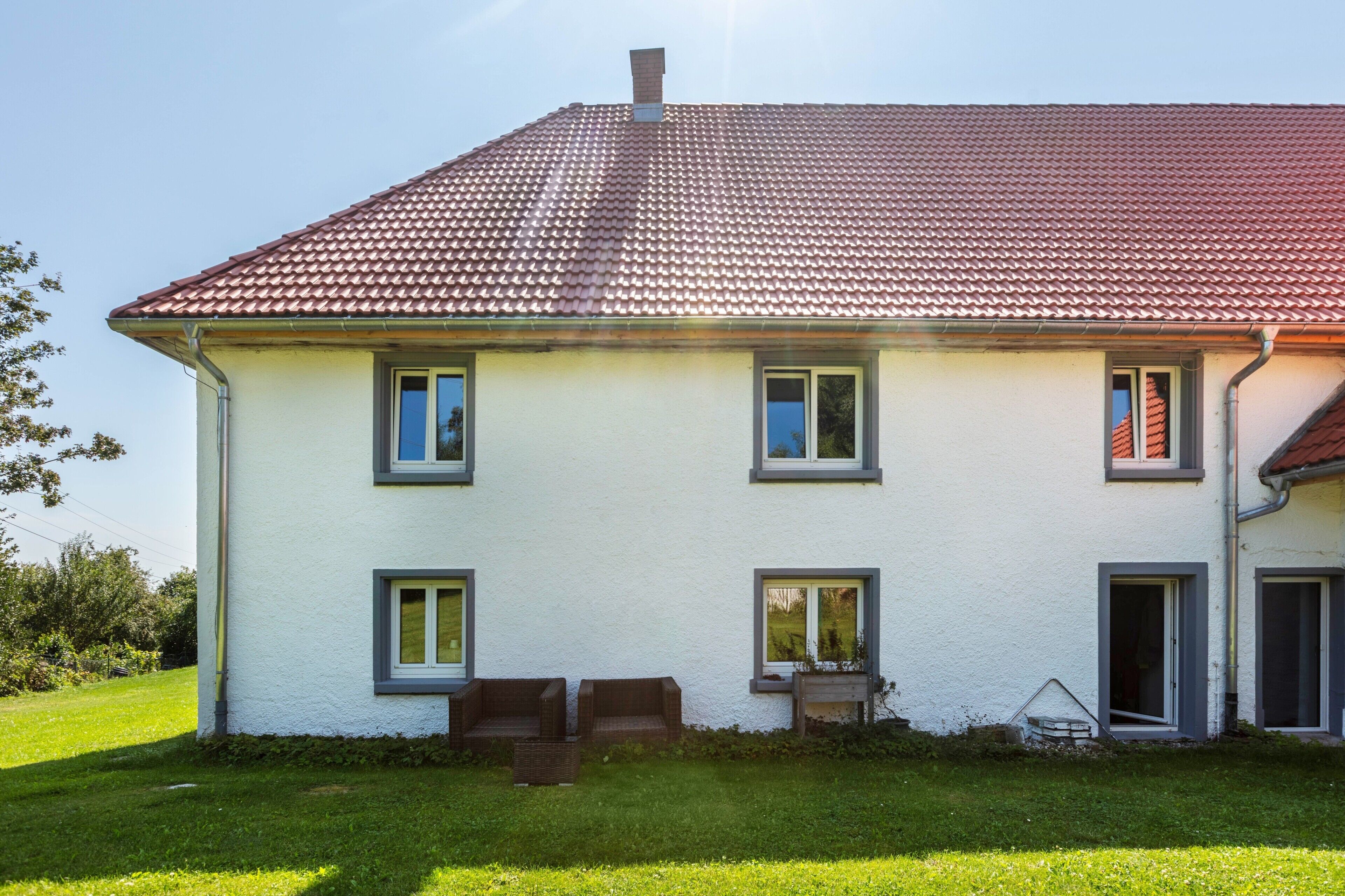 A two-story house with a red-tiled roof, white exterior walls, and a small front porch with two chairs.