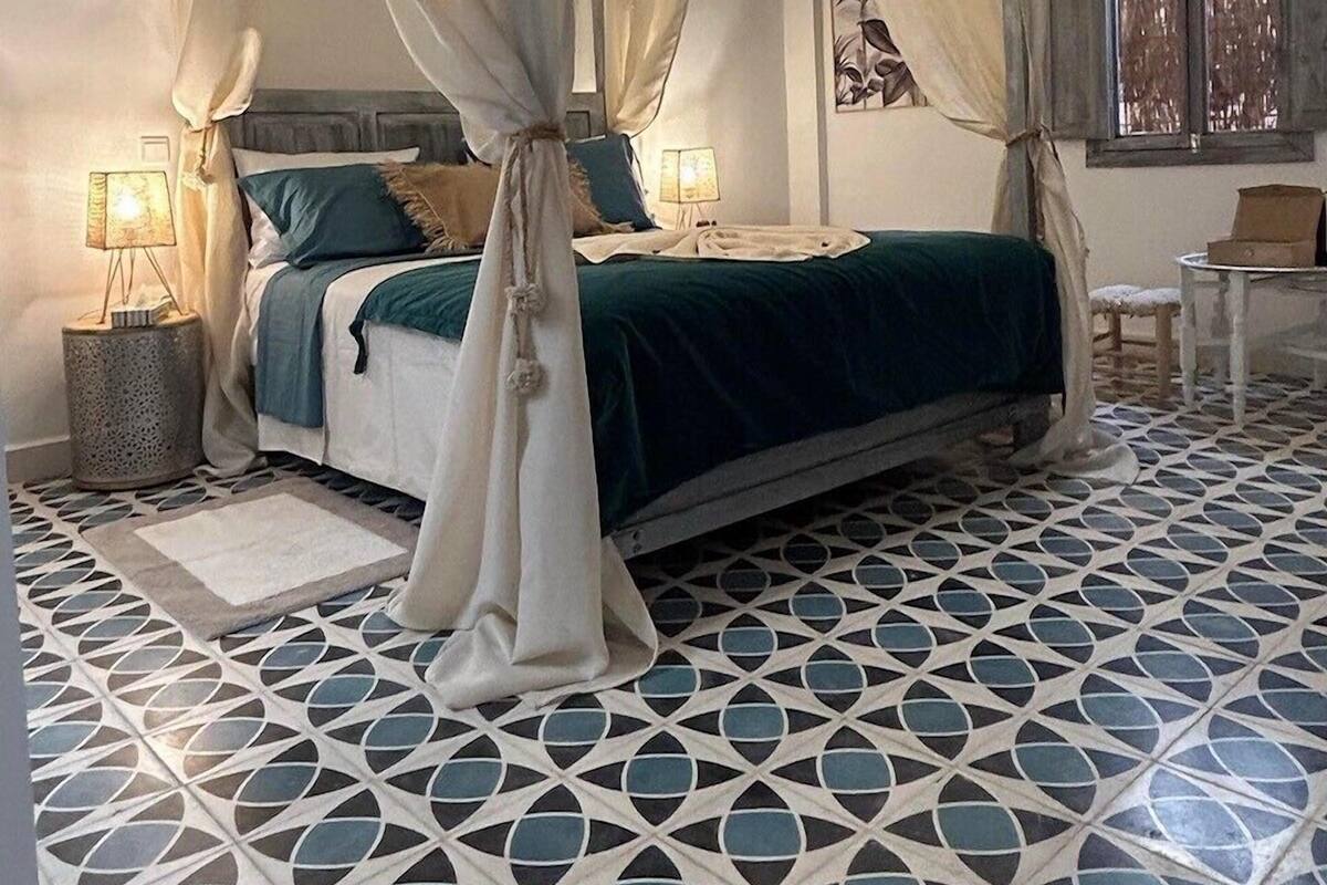 A bedroom with a four-poster canopy bed, a patterned floor, and a small table with a chair.