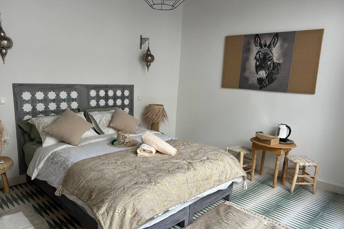 A neatly arranged bedroom with a bed, bedside tables, a small stool, a wall-mounted lamp, and a framed picture.
