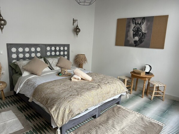 A neatly arranged bedroom with a bed, bedside tables, a small stool, a wall-mounted lamp, and a framed picture.