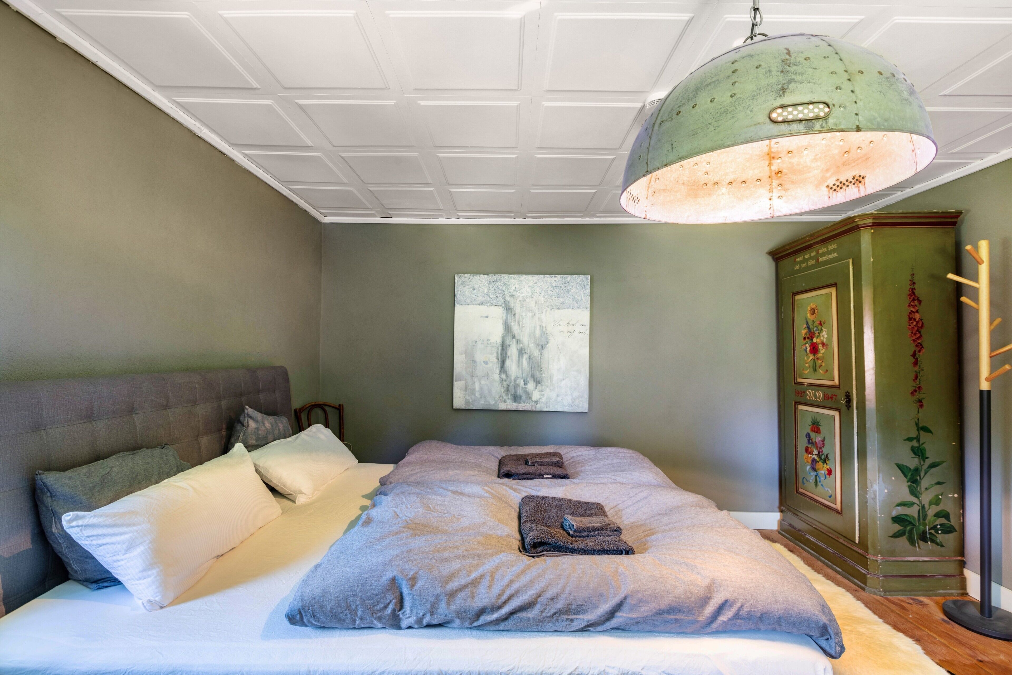 A bedroom with a bed, pillows, a hanging light fixture, a painting, and a cabinet.