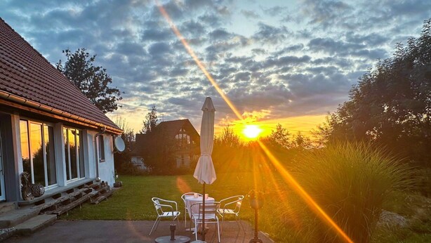 Een terras met tuinmeubilair, een tafel en een parasol, met een huis en bomen op de achtergrond.
