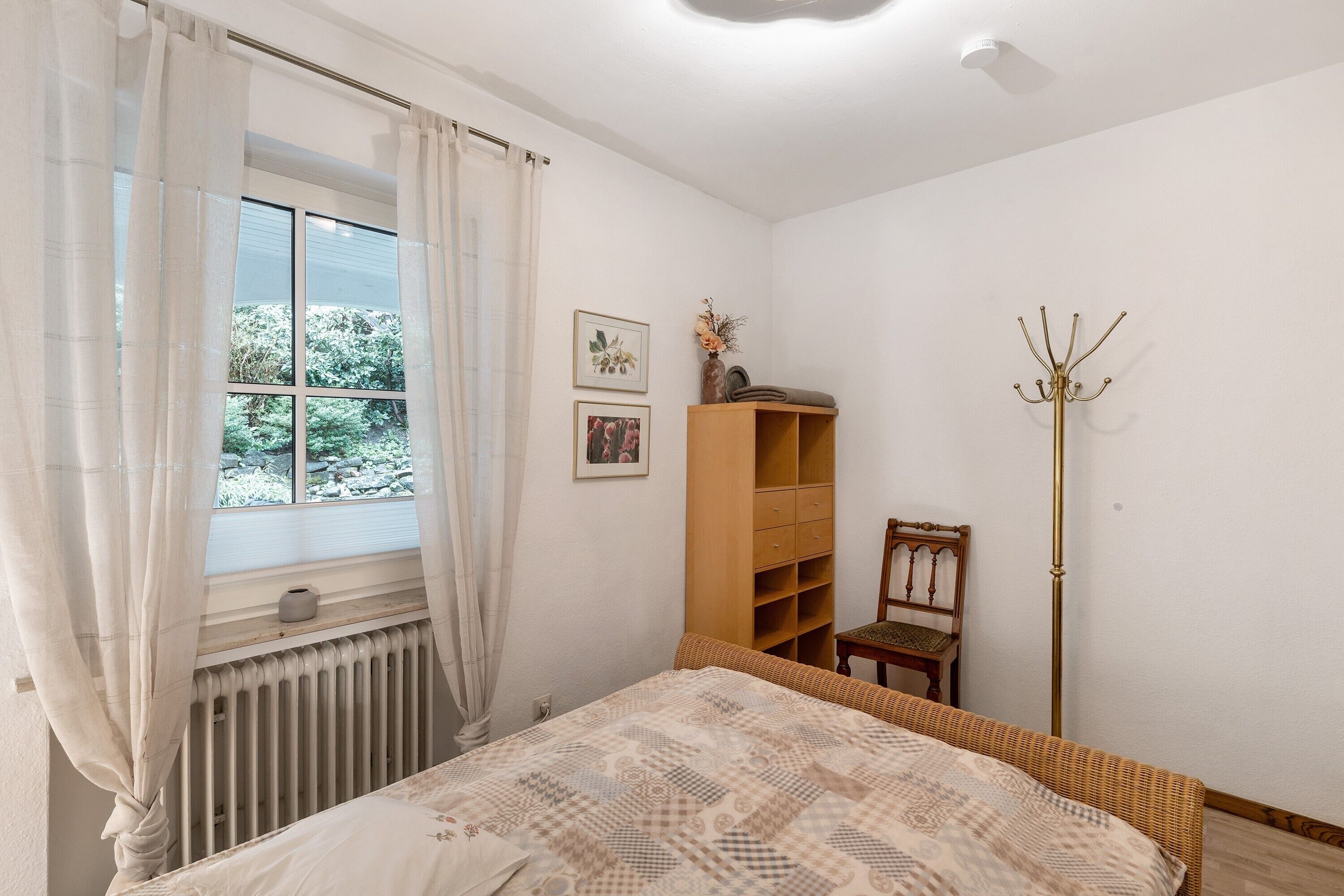 Een slaapkamer met een bed, een radiator, een raam met gordijnen, een houten plank, een stoel en een kapstok.