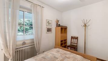 Een slaapkamer met een bed, een radiator, een raam met gordijnen, een houten plank, een stoel en een kapstok.
