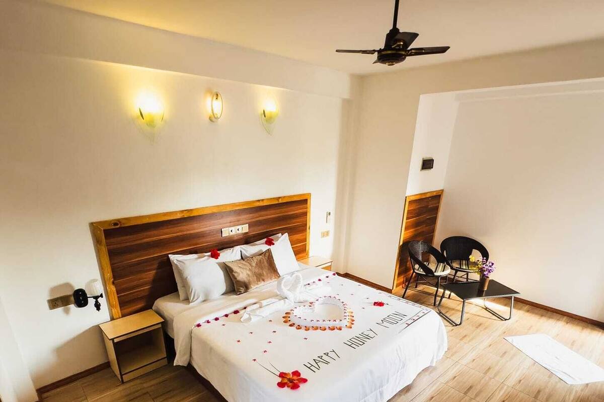 Una habitación de hotel con suelo de madera, una cama con cubrecama blanco, una mesita de noche, una silla y un ventilador de techo.