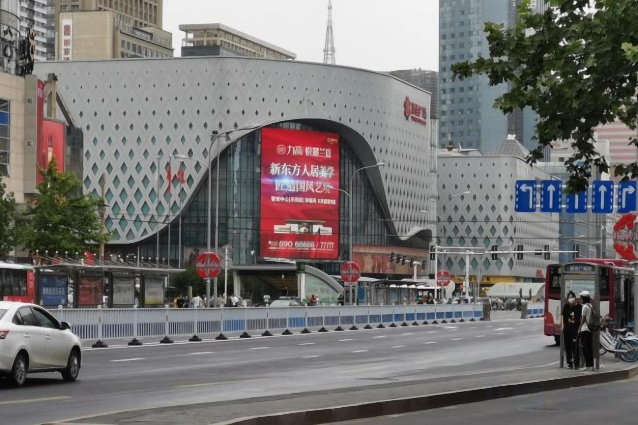 Ein modernes Gebäude mit geschwungener Fassade, einer großen digitalen Werbung und einer Bushaltestelle mit Personen.