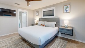 A bedroom with a bed, a ceiling fan, a TV, a window with blinds, and a nightstand with a lamp.